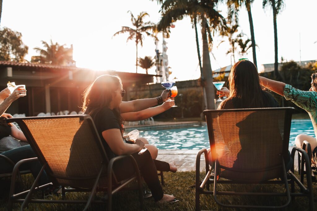 Group of friends in casual clothing drinking alcoholic beverages while resting on deckchairs on hotel territory by swimming pool at sunset